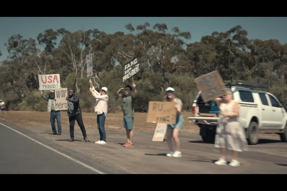 A group of people standing along a rural roadside holding handmade protest signs, with a white ute parked nearby and Australian gum trees in the background