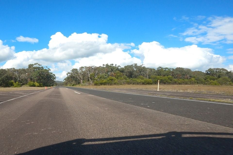 A view from inside a vehicle looking out over an open two-lane road lined with eucalyptus trees under a blue sky with scattered clouds in what appears to be a rural Australian setting.