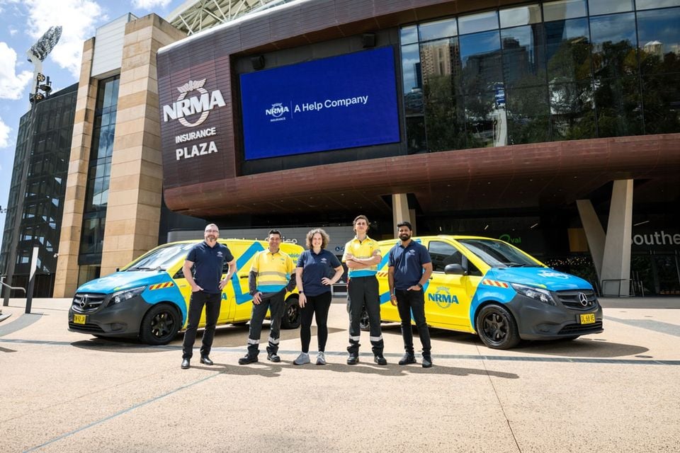 Five NRMA roadside staff standing between two yellow and blue Mercedes-Benz Vito patrol vans outside NRMA Insurance Plaza at Adelaide Oval