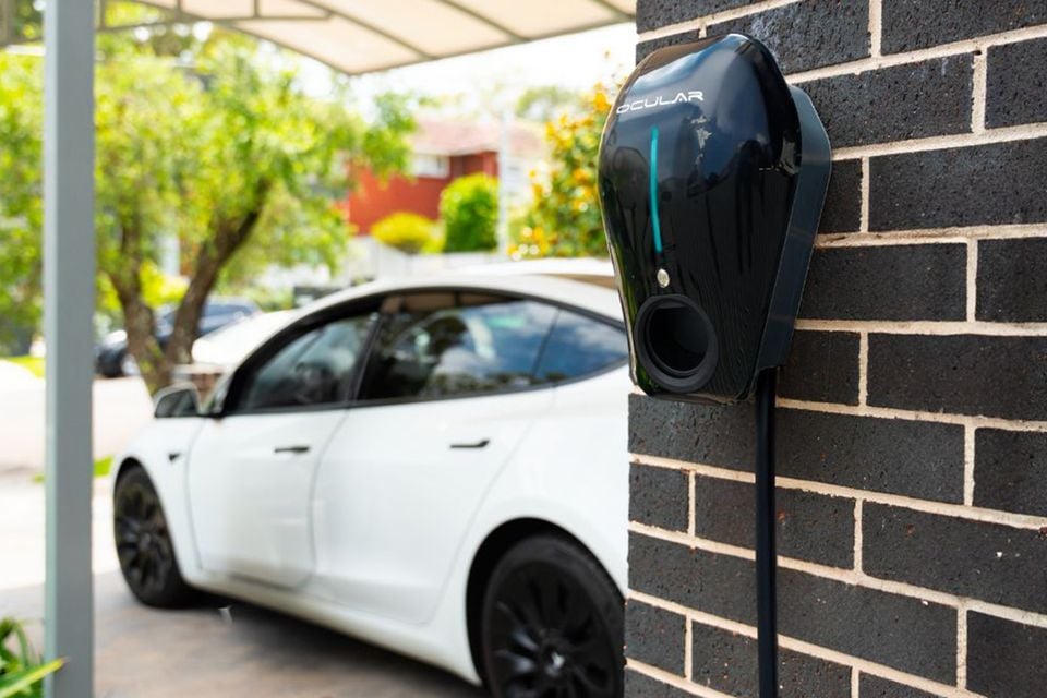 An Ocular IQ home EV charger wall-mounted on a brick wall, with a white Tesla sedan parked in the background under a carport.