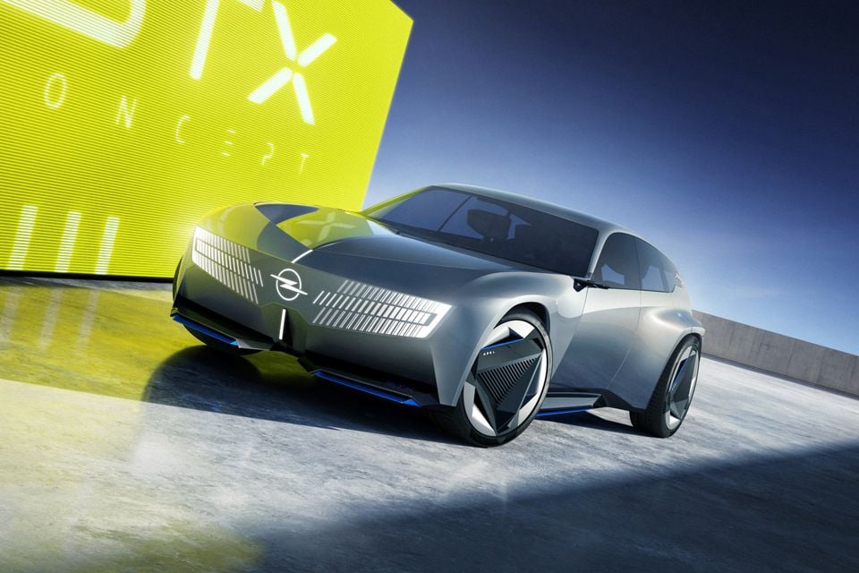 Um SUV conceito Opel STX cinza visto da parte frontal, com barras de luz iluminadas, estacionado em frente a um display STX Concept amarelo