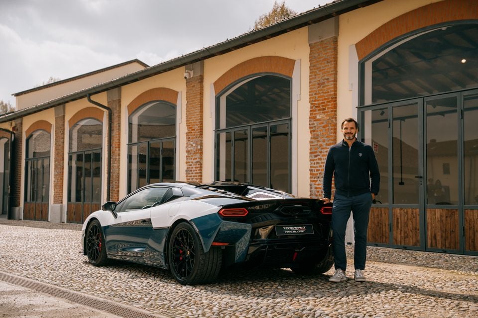 A man stands beside a blue and white Lamborghini Temerario Tricolore supercar parked outside a brick building, viewed from the rear quarter.