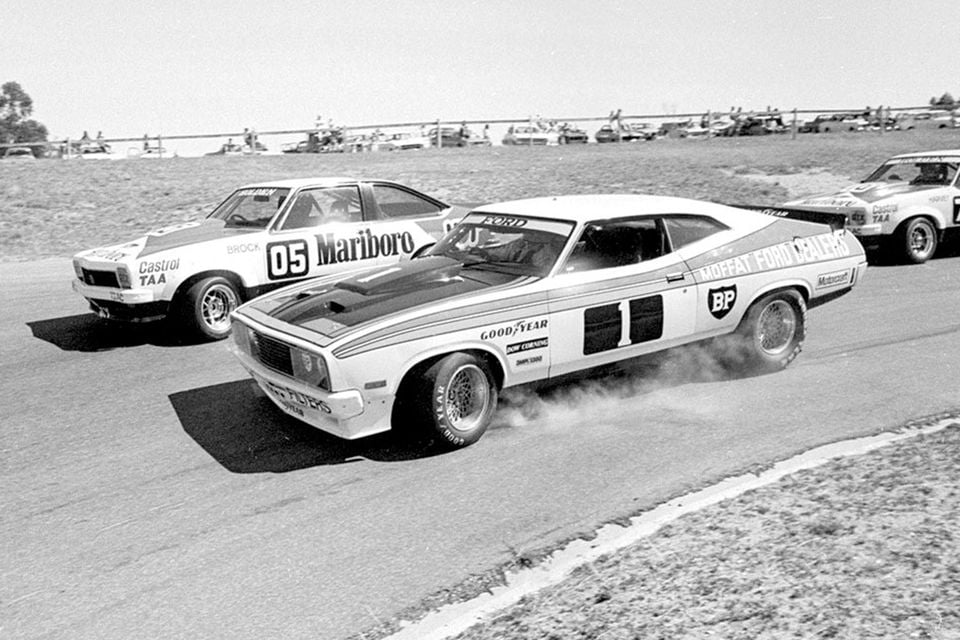 A black-and-white action photo of Allan Moffat's No. 1 Moffat Ford Dealers Ford Falcon racing car leading Peter Brock's No. 05 Marlboro Holden Torana on a race circuit, with tyre smoke visible from the Ford.