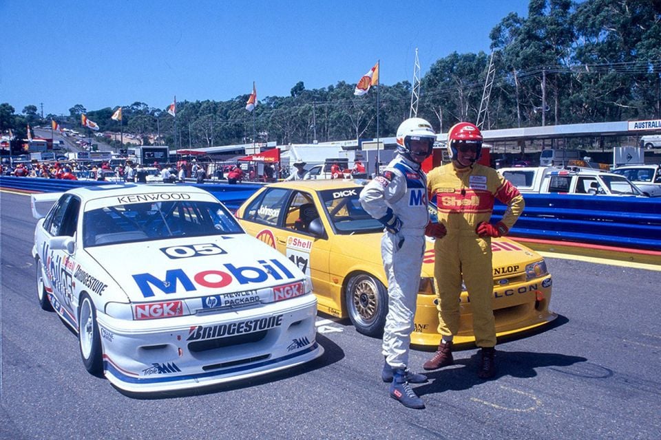 Two racing drivers in full race suits and helmets stand between a white Mobil-liveried Holden and a yellow Shell-liveried Ford Falcon at what appears to be Bathurst circuit in 1993