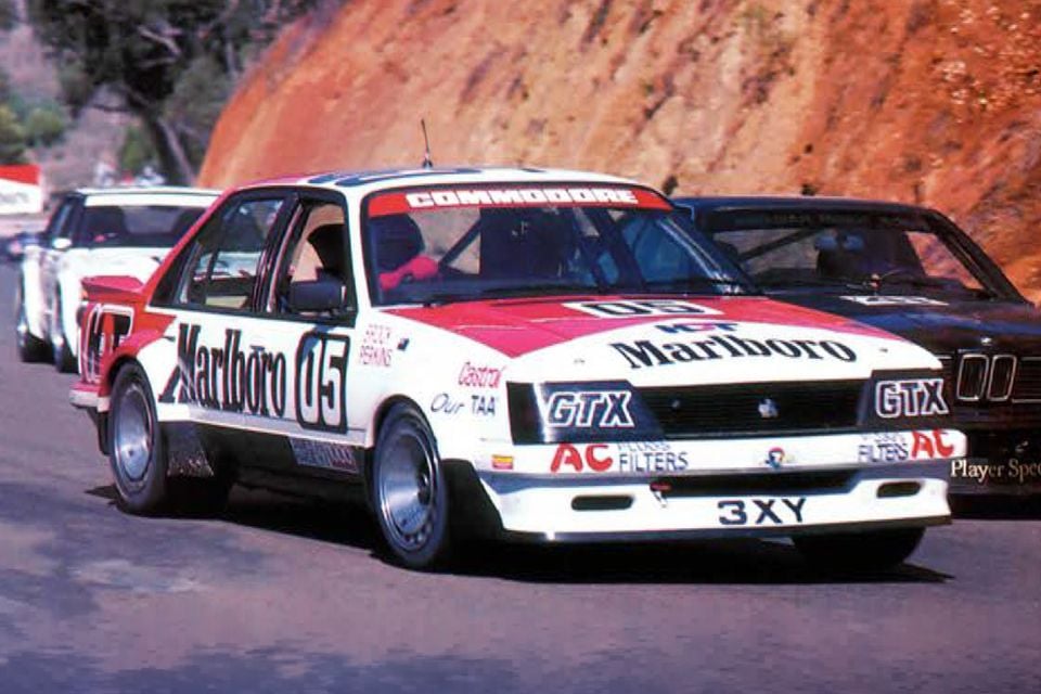 A Holden VH Commodore race car with Marlboro livery, number 05, driven by Peter Brock and Larry Perkins on circuit at Bathurst in 1982, seen from the front-side with other race cars close behind
