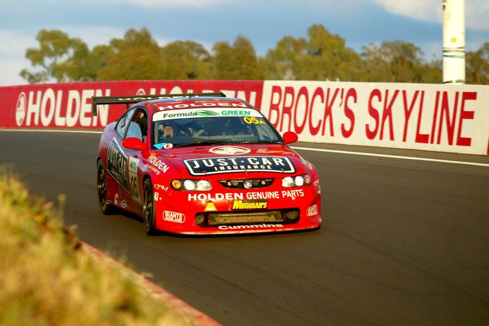 A red Holden Monaro V8 Supercar race car with Just Car Insurance and Holden Genuine Parts livery competing at Bathurst, passing the Brock's Skyline signage on the circuit