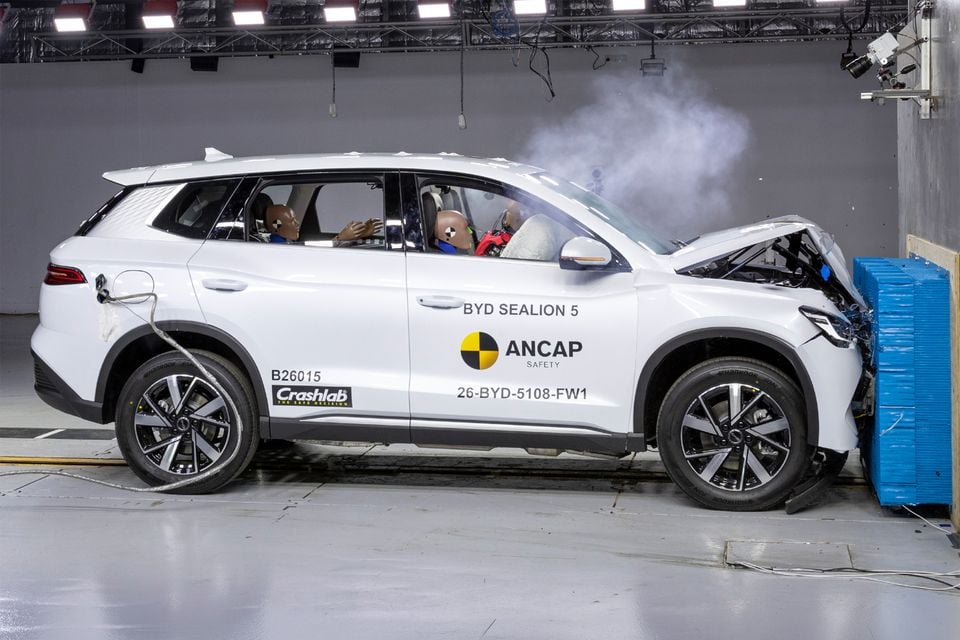 A white BYD Sealion 5 SUV with crash test dummies inside undergoes an ANCAP frontal offset crash test, its front end crumpled against a barrier.
