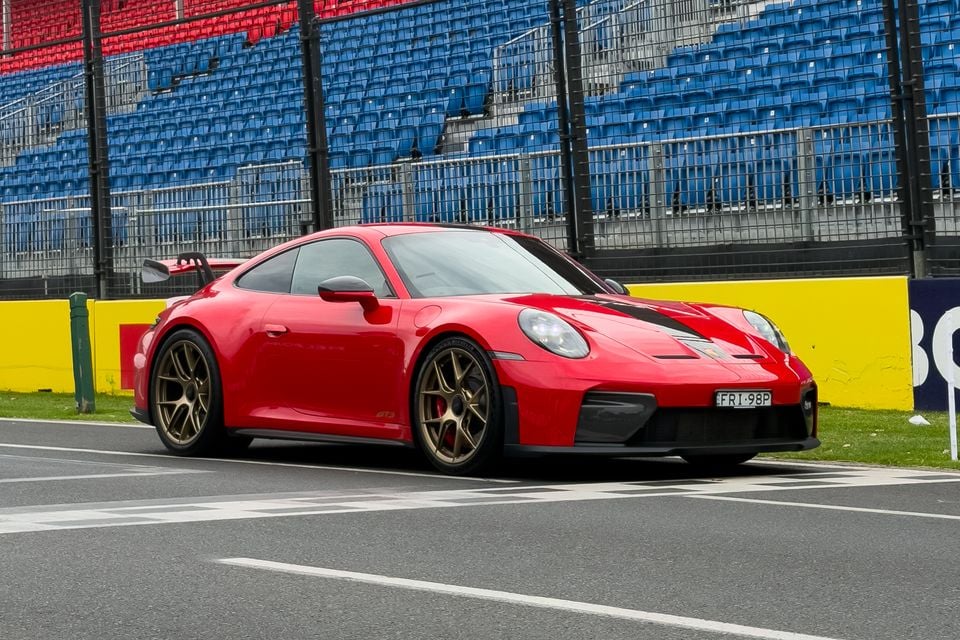 A red Porsche 911 GT3 with Weissach Package coupe with gold wheels parked at a motorsport circuit, seen from the front-side angle with grandstand seating in the background