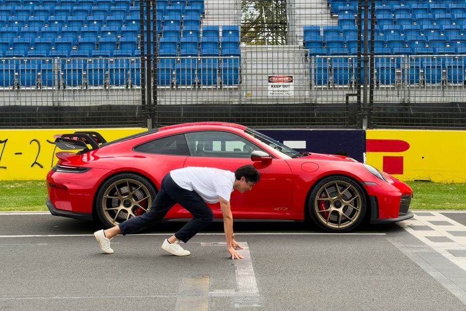 A red Porsche 911 GT3 parked on a racing circuit pit lane, with a person crouching at the starting line in front of it and empty grandstands visible in the background