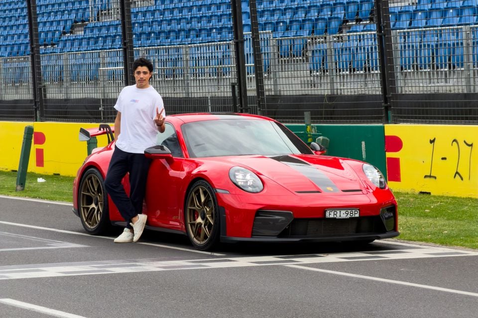 A red Porsche 911 GT3 with Weissach Package parked at a racing circuit, with a young man standing beside it giving a peace sign.