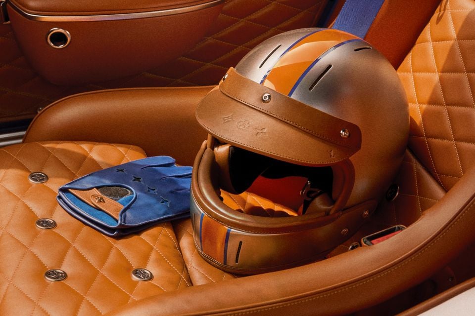 A luxury racing helmet with tan leather trim and orange and blue detailing, along with matching blue leather driving gloves, resting on the quilted tan leather seat of a Porsche 911 reimagined by Singer x Louis Vuitton.