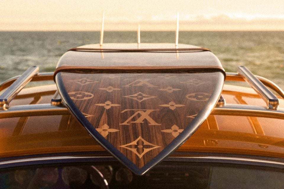 A Louis Vuitton monogram-patterned wooden surfboard mounted on the roof rack of a Porsche 911 reimagined by Singer, with a golden-orange car body and ocean visible in the background.