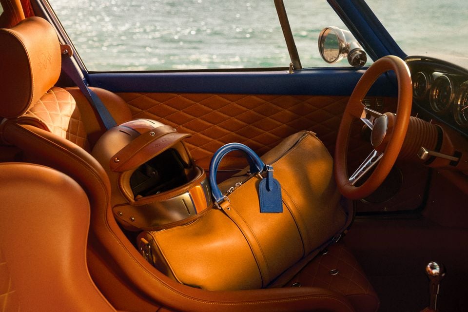 The tan leather interior of a classic Porsche 911 reimagined by Singer, featuring diamond-stitched seats, a wooden-rimmed steering wheel, a racing helmet and a Louis Vuitton tan leather duffle bag on the passenger seat, with water visible through the window.