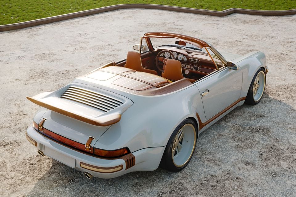 A pale grey custom Porsche 911 cabriolet, reimagined by Singer and Louis Vuitton, seen from the rear three-quarter angle, featuring gold and tan leather accents throughout, parked on a gravel surface