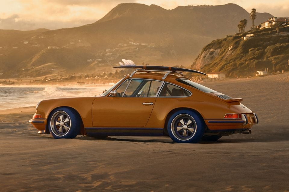 A golden-orange classic Porsche 911 coupe with a surfboard on the roof rack, parked on a sandy beach at sunset with mountains and ocean in the background