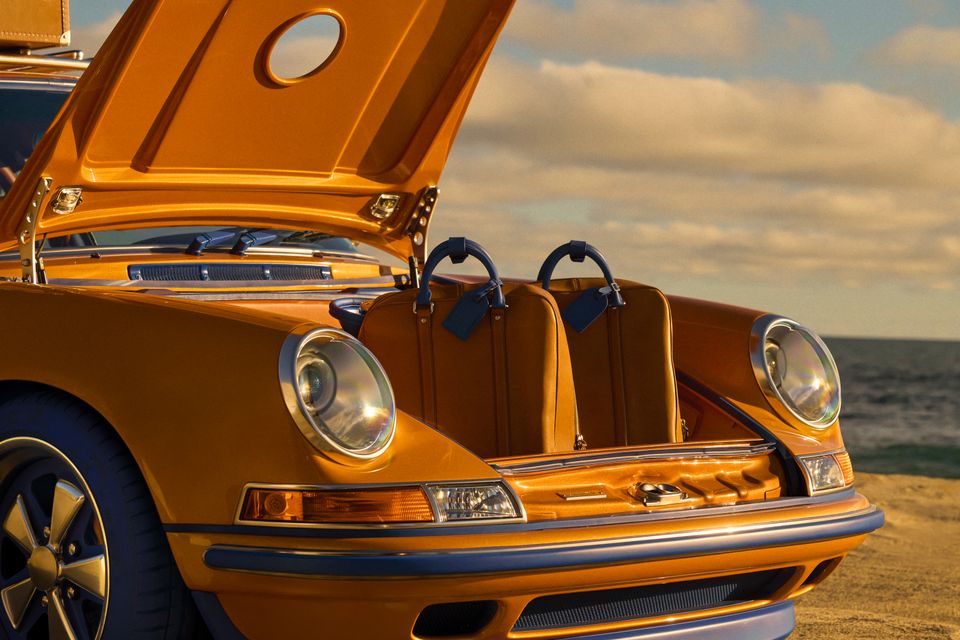 The open front trunk of a gold Singer-reimagined Porsche 911 fitted with custom matching luggage, parked near a beach at sunset