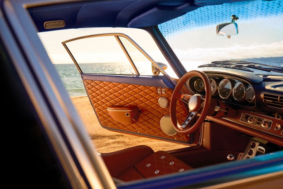 Interior of a custom Porsche 911 reimagined by Singer and Louis Vuitton, showing tan diamond-quilted leather door panel, steering wheel, and gauges, photographed at a beachside location
