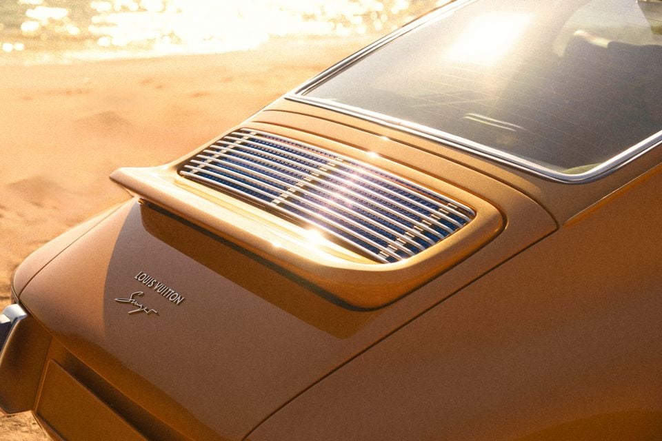 Close-up of the rear engine lid grille on a Singer-reimagined Porsche 911 in gold, badged with Louis Vuitton and Singer logos, bathed in warm sunlight