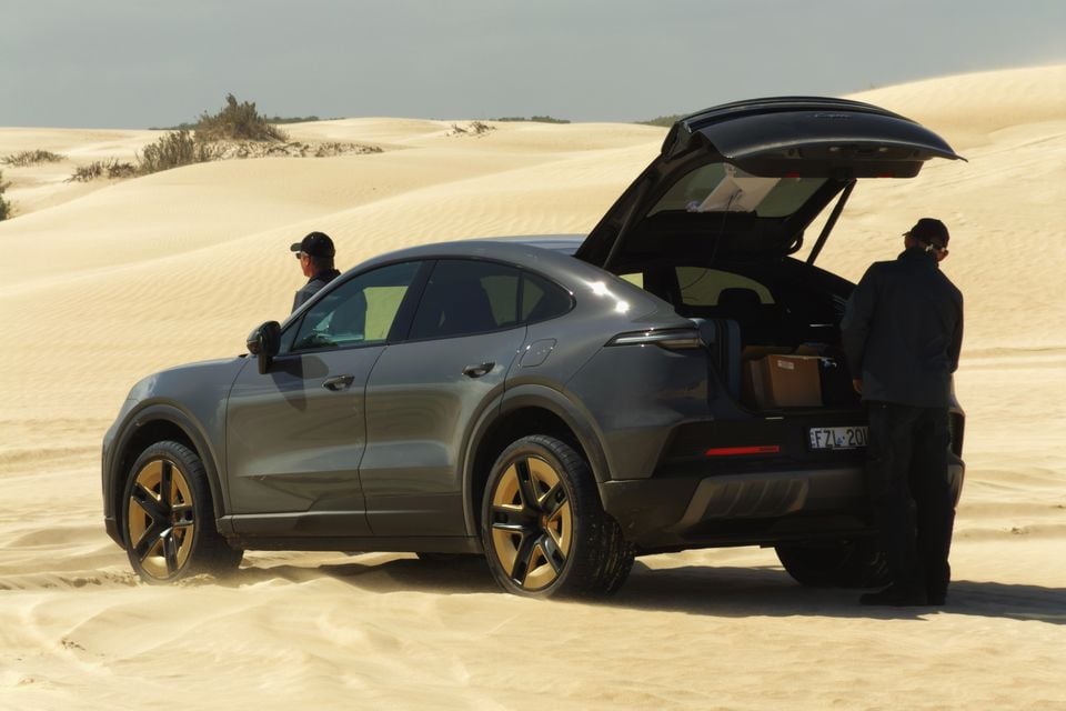 A grey Porsche Cayenne Coupe SUV parked on sand dunes with its boot open, with two people standing nearby