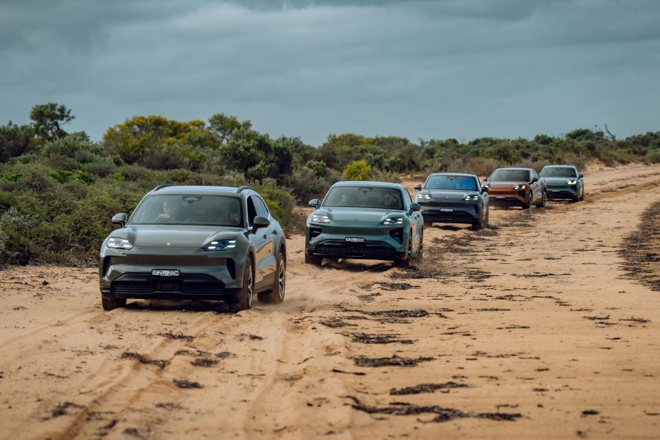 A convoy of Porsche Cayenne Electric SUVs driving along a sandy dirt track through Australian coastal scrubland, seen from the front.