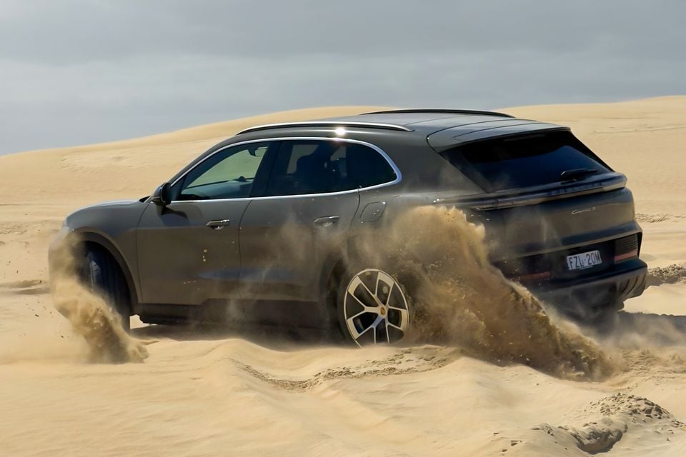 A Porsche Cayenne S SUV driving through sand dunes, kicking up a large cloud of sand, seen from the rear three-quarter angle