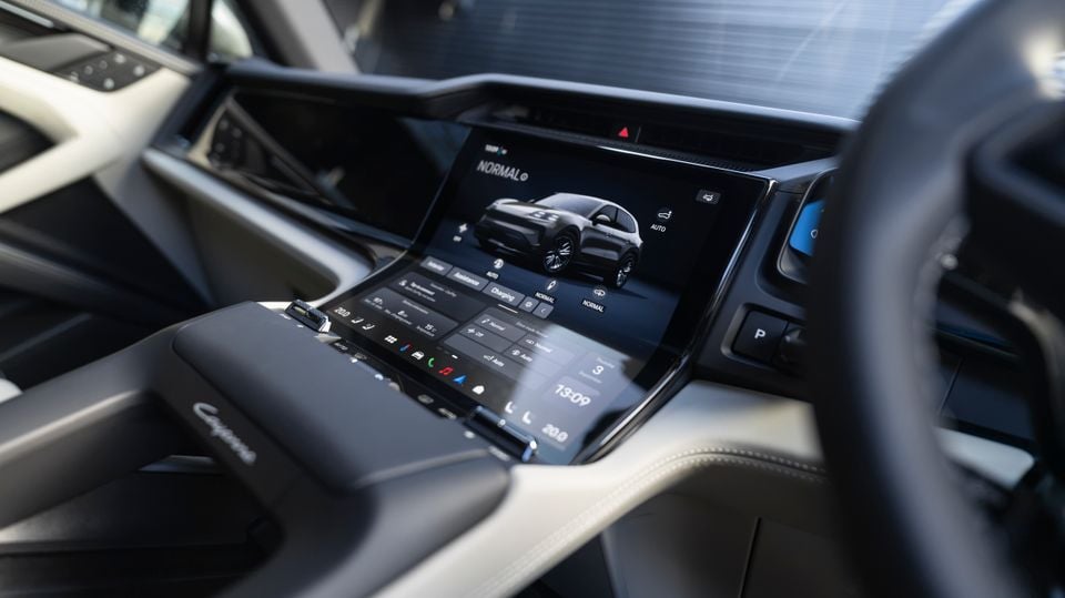 Interior view of a Porsche Cayenne S Electric showing the curved infotainment touchscreen displaying drive mode settings, alongside a secondary control panel on the centre console