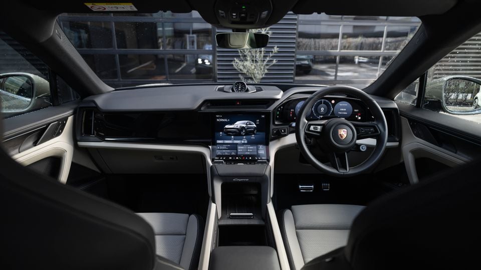 Interior of a Porsche Cayenne showing the dashboard, large central touchscreen infotainment system, digital instrument cluster, and steering wheel with Porsche crest, shot from the driver's perspective in right-hand drive