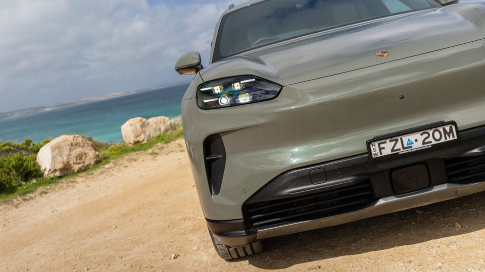 Close-up of the front end of a green Porsche Cayenne S Electric SUV, parked on a dirt coastal lookout with turquoise ocean in the background