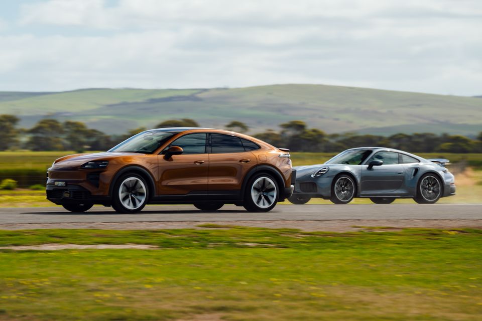 An orange Porsche Cayenne Turbo Coupe electric SUV driving alongside a grey Porsche 911 Turbo coupe on a country road, seen from the side.