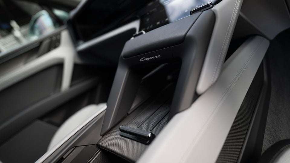 Interior of a Porsche Cayenne showing the centre console armrest with 'Cayenne' branding, carbon fibre trim, and light leather seats