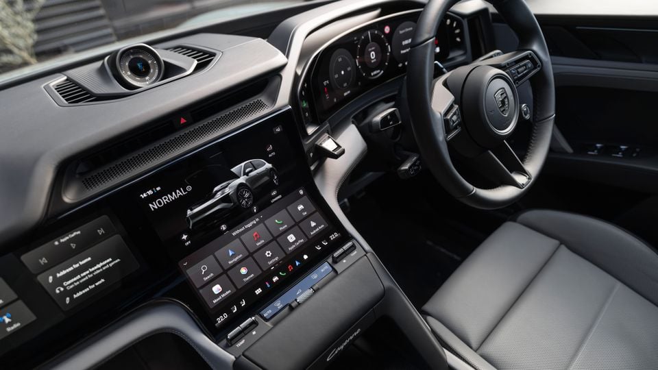 Interior of a Porsche Cayenne Turbo E-Hybrid showing the large central touchscreen infotainment system displaying Normal drive mode, with the steering wheel, instrument cluster, and front passenger seat visible