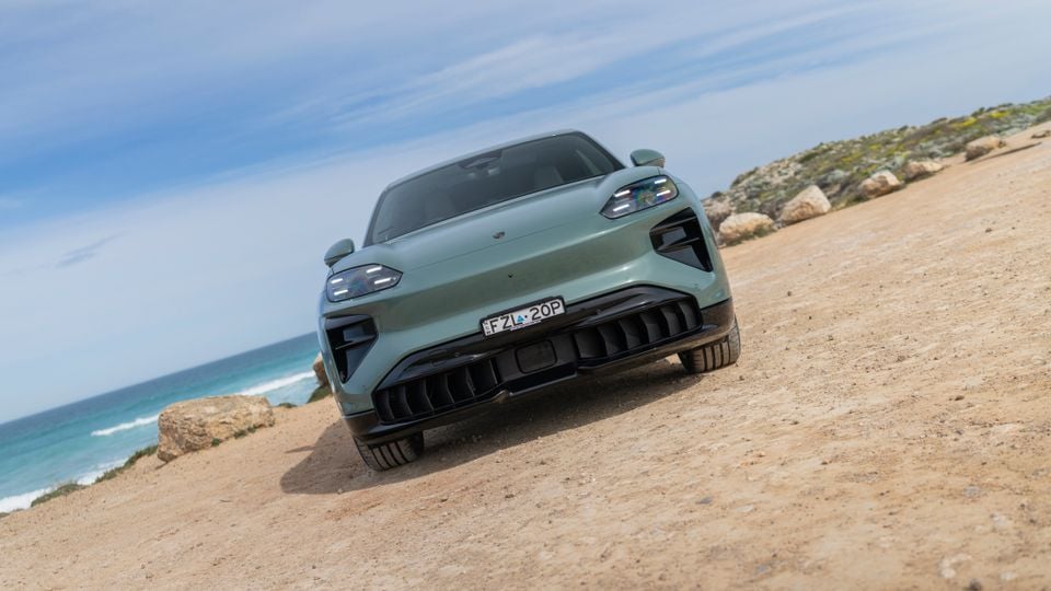 A green Porsche Cayenne Turbo Electric SUV seen from the front, parked on a sandy coastal track with the ocean in the background.