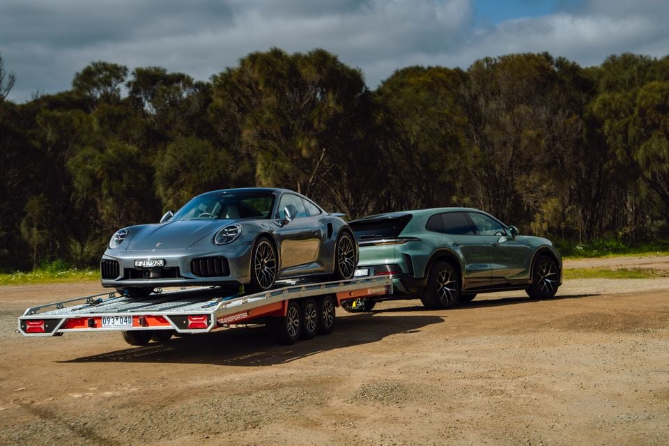 A grey Porsche 911 loaded on a car transport trailer being towed by a green Porsche Cayenne Turbo E-Hybrid on a dirt road in Australia