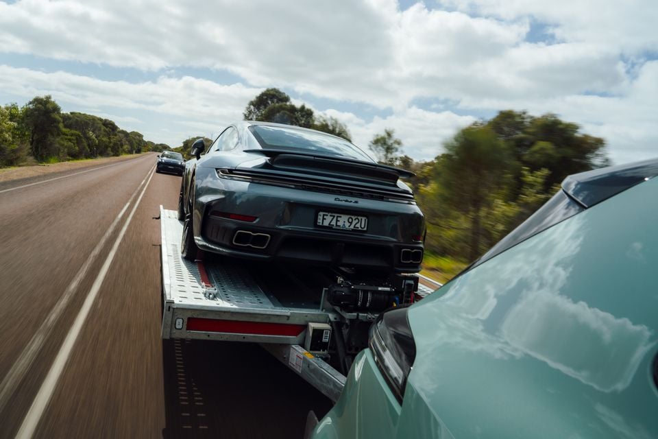 A dark Porsche 911 Turbo S loaded on a flatbed trailer being towed along an Australian highway, seen from the rear, with a camera rig mounted beneath it
