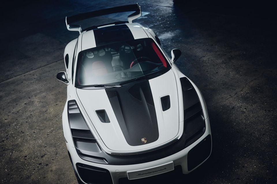 A white Porsche 911 sports car with carbon fibre accents and a large rear wing, seen from a high front angle in a darkened studio setting