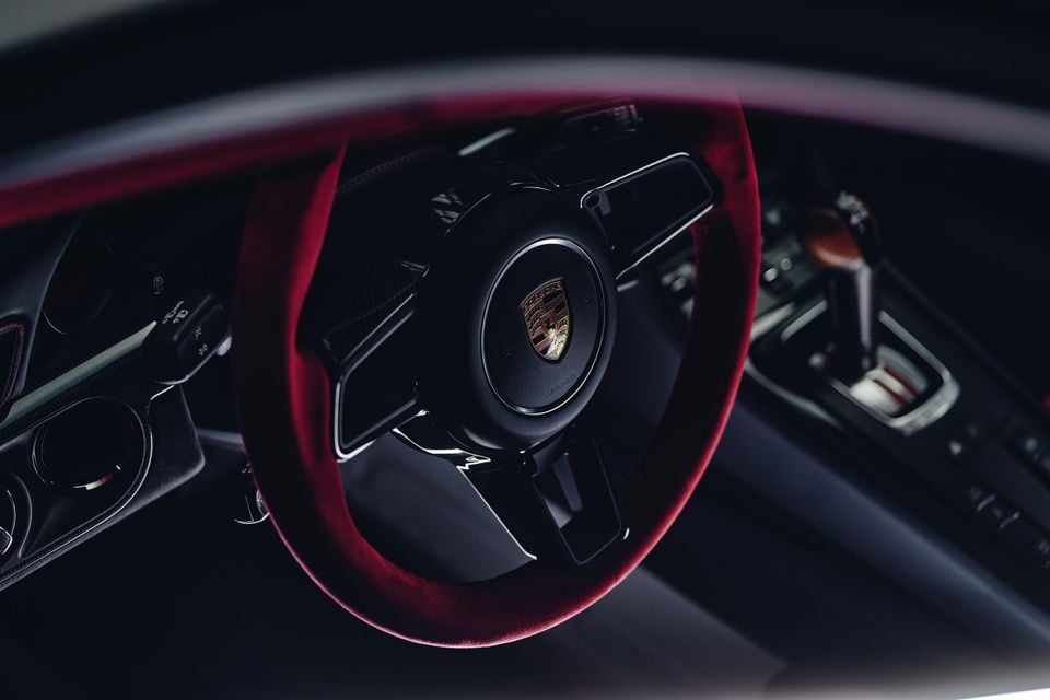 Close-up of a Porsche steering wheel with a red alcantara rim and Porsche crest badge, with a gear selector visible in the background