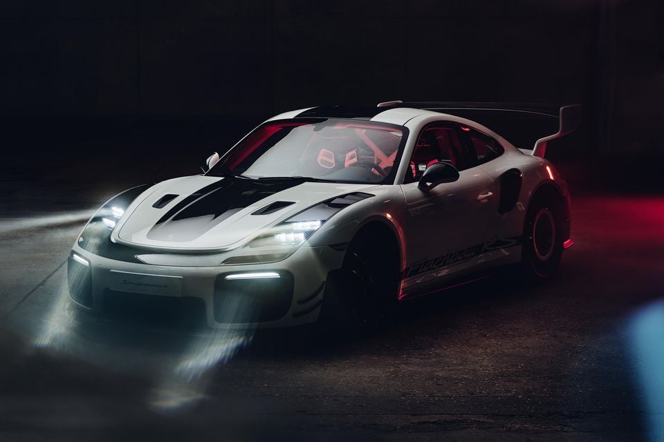 A white Porsche 911 GT2 RS sports car with black hood stripes, large rear wing, and red interior lighting, seen from the front-side in a dark studio setting