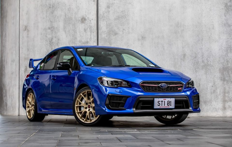 A blue Subaru WRX STI Saigo special edition sedan with gold wheels, seen from the front-side, parked against a concrete wall with a 'STI 01 – Last of a Generation' number plate.