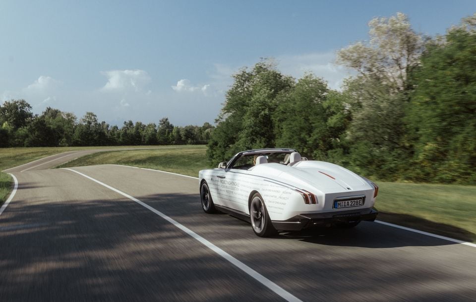 A white Rolls-Royce Droptail convertible, labelled "Project Nightingale," is driven around a curving country road, seen from the rear quarter.