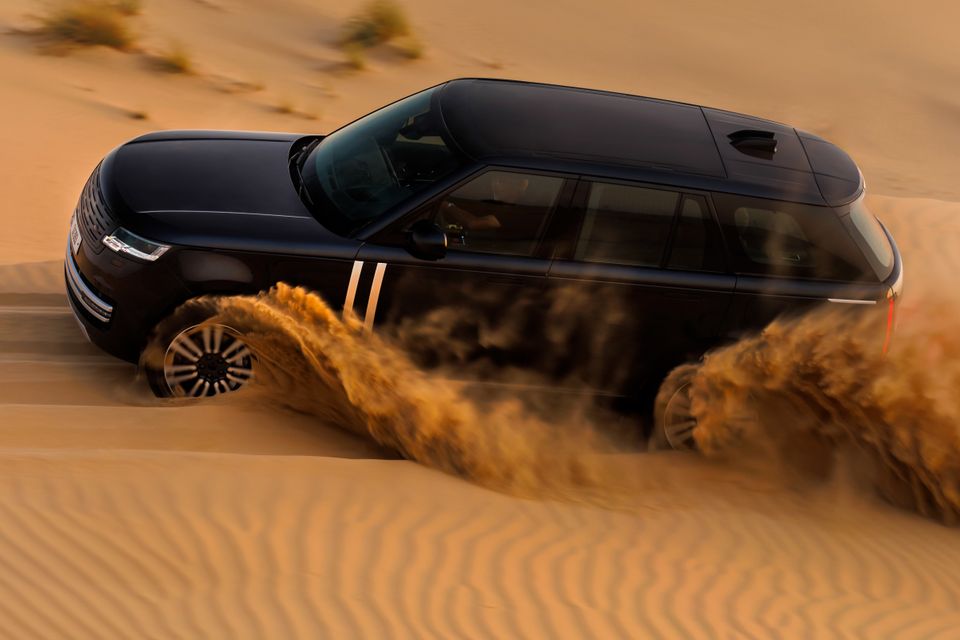 A dark blue Range Rover SUV driving through sand dunes in a desert, kicking up large clouds of sand