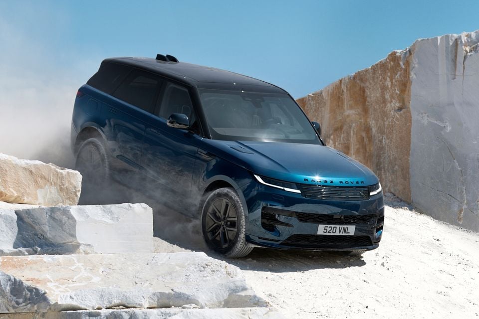 A dark blue Range Rover Sport SUV navigates a dusty quarry, seen from the front quarter with a stone wall in the background.
