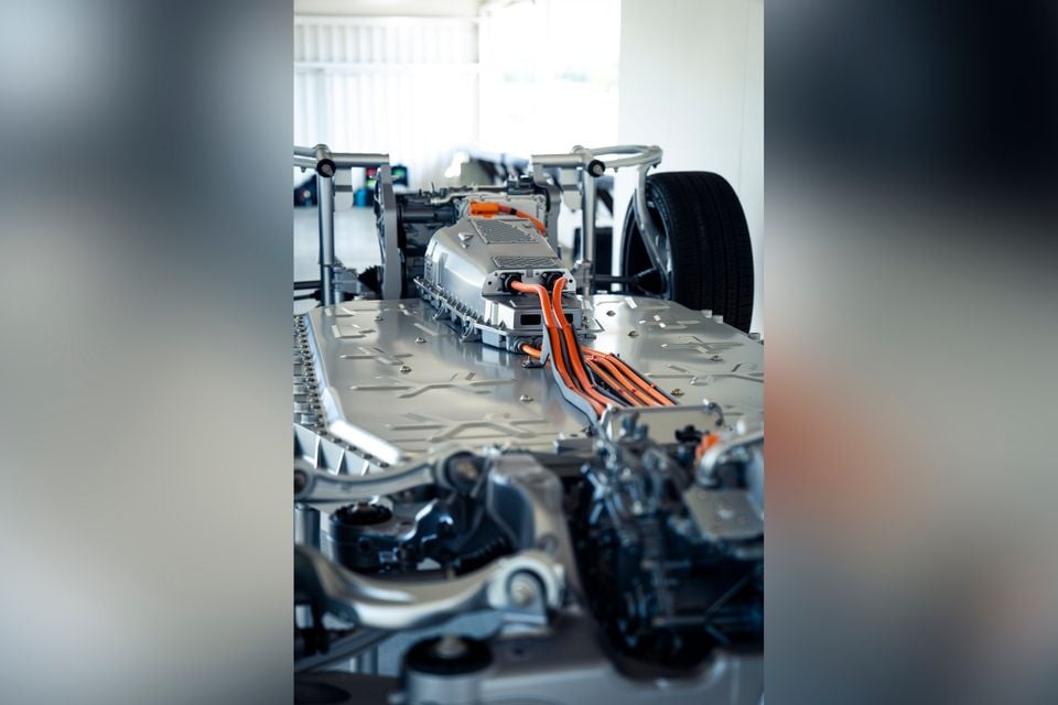 Close-up of an electric vehicle chassis showing orange high-voltage cables running along the battery pack floor, with suspension components and a tyre visible in the background