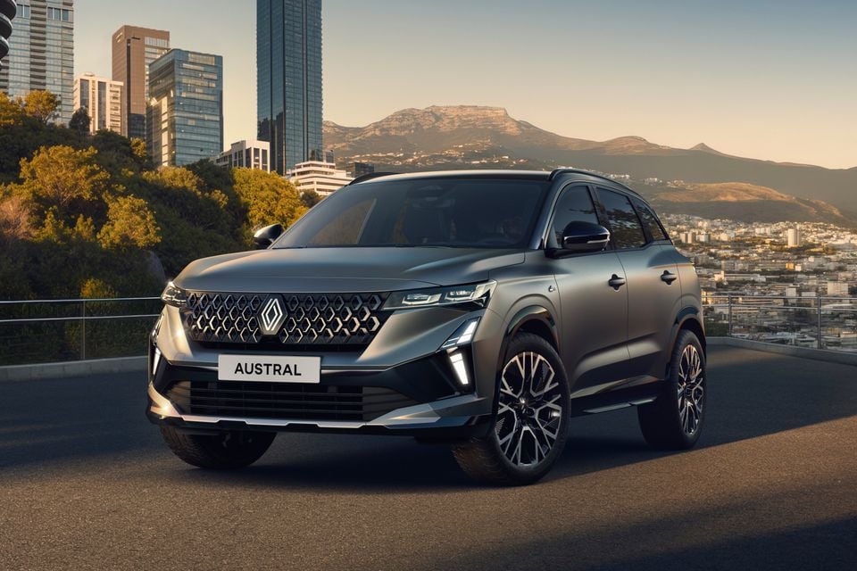 A grey Renault Austral SUV seen from the front-side angle, parked on an elevated road with a city skyline and mountains in the background.