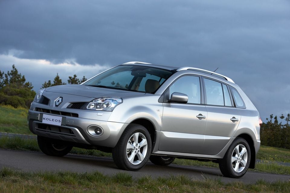A silver Renault Koleos SUV seen from the front-side angle, parked on a road with trees and cloudy sky in the background.