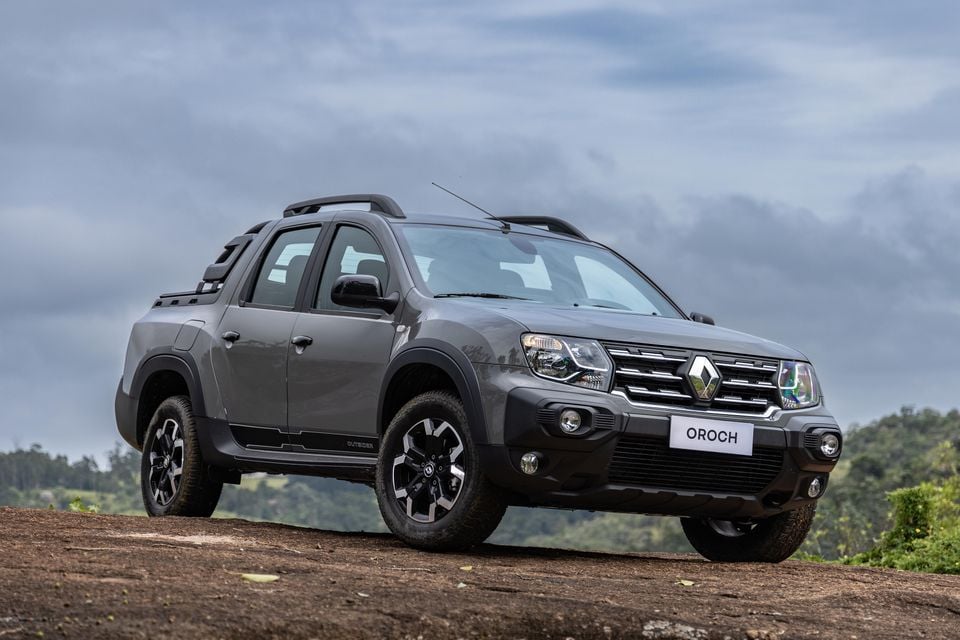 A grey Renault Oroch dual-cab ute seen from the front-left side, parked on a dirt rise with a cloudy sky and green hills in the background.