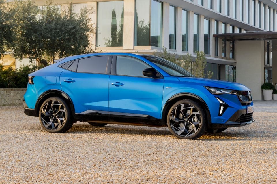 A blue Renault Rafale E-Tech 4x4 SUV coupe seen from the side, parked on a gravel area outside a modern building