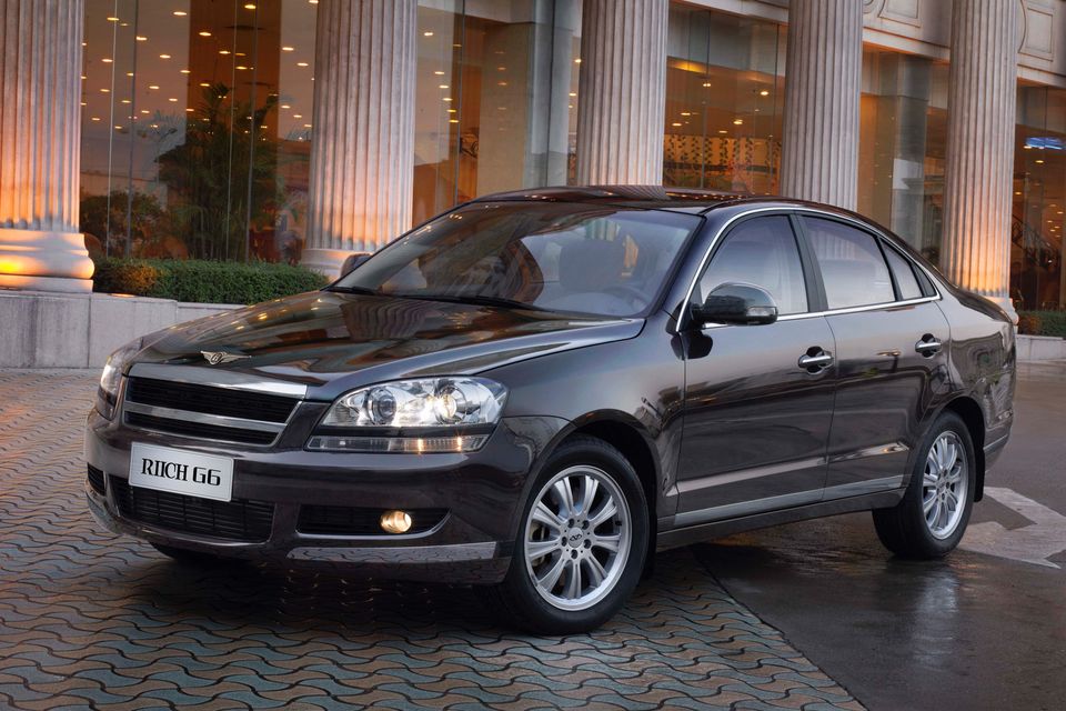 A dark grey Riich G6 sedan parked in front of a building with large columns, seen from the front-left angle.
