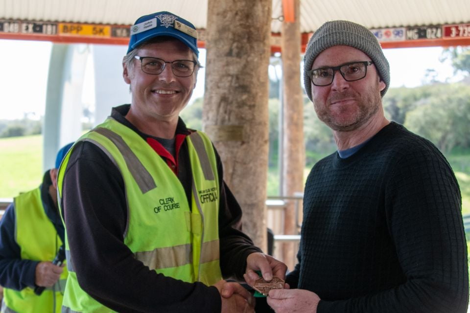 A clerk of course in a high-visibility vest shakes hands with a man in a beanie while presenting a medal at the Rob Roy Hillclimb