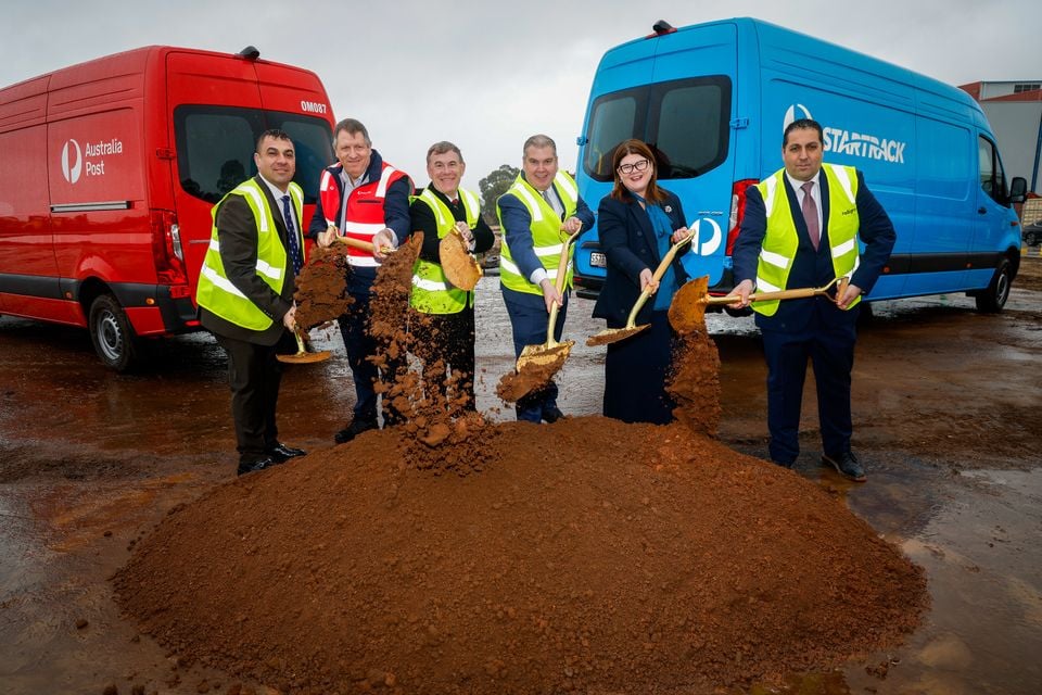 Six people in high-visibility vests perform a ceremonial sod-turning with gold shovels at a construction site, flanked by an Australia Post red van and a blue StarTrack van.