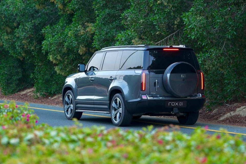 A grey Rox 01 SUV with a rear-mounted spare wheel driving along a tree-lined road, seen from the rear